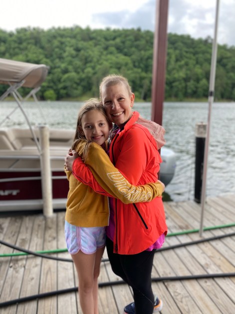Tween girl and bald mother hugging by a pontoon boat at a lake